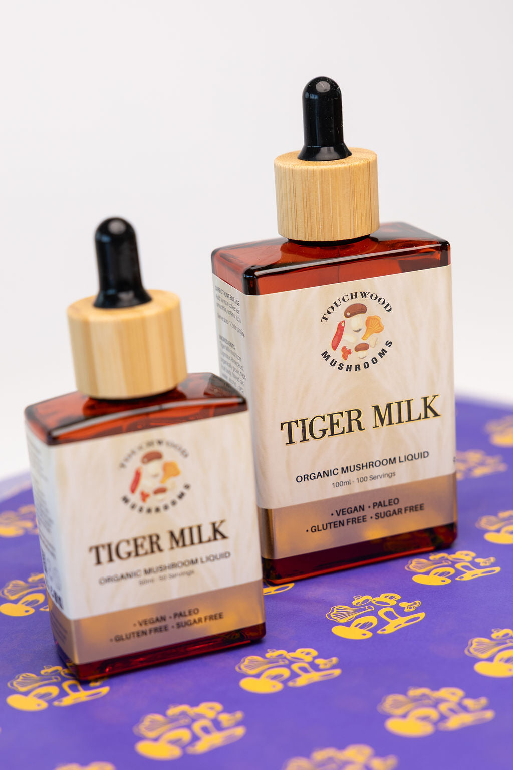 Two bottles of Tiger Milk organic mushroom liquid on a purple background with yellow tiger paw stamped print.